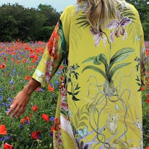 Op de afbeelding: Een lange, vloeiende gele kimono met een botanische print met orchidee&euml;n, vlinders en andere tropische elementen. Het ontwerp van de kimono bevat een mix van bloemen- en dierlijke illustraties. De achtergrond toont een veld met rode klaprozen en korenbloemen.