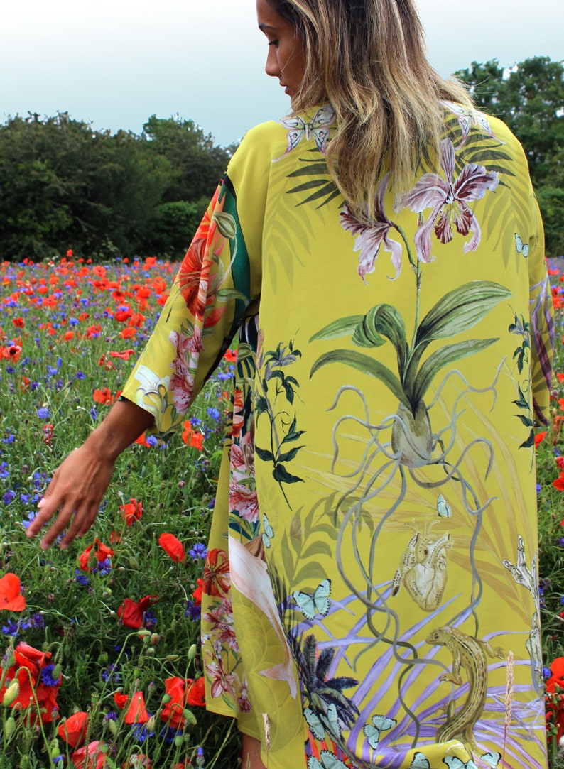 Op de afbeelding: Een lange, vloeiende gele kimono met een botanische print met orchidee&euml;n, vlinders en andere tropische elementen. Het ontwerp van de kimono bevat een mix van bloemen- en dierlijke illustraties. De achtergrond toont een veld met rode klaprozen en korenbloemen.