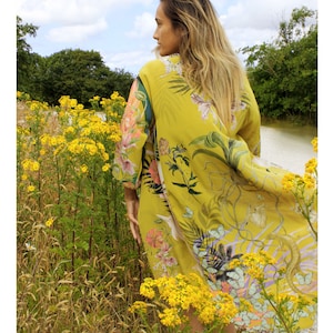 Op de afbeelding: Een gele kimono-achtige jurk met een botanische print van bloemen en vlinders. De jurk wordt buiten gedragen, te midden van gele wilde bloemen, bomen en een waterpartij. De stof is licht en luchtig.