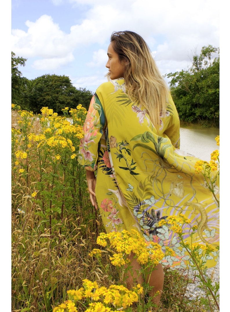 Op de afbeelding: Een gele kimono-achtige jurk met een botanische print van bloemen en vlinders. De jurk wordt buiten gedragen, te midden van gele wilde bloemen, bomen en een waterpartij. De stof is licht en luchtig.
