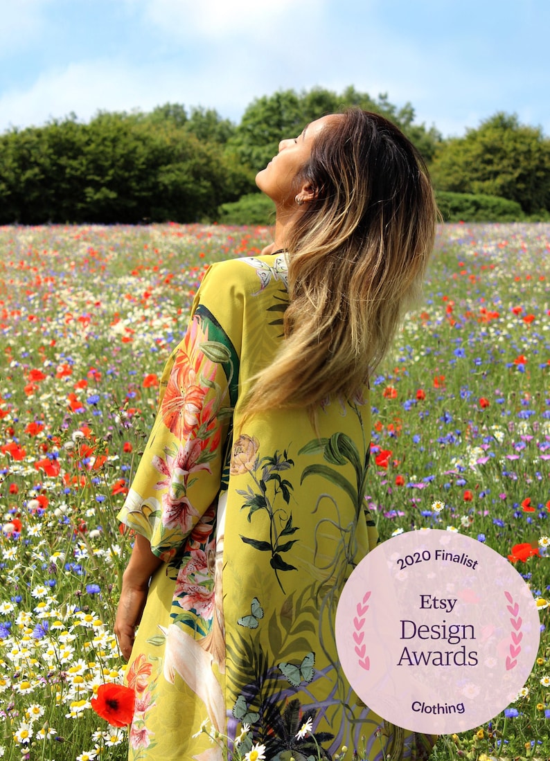 Op de afbeelding: Een vrouw draagt een gele bloemenkimono in een veld met wilde bloemen. De kimono heeft een levendige bloemenprint met rode, roze en groene accenten. De afbeelding bevat een 2020 Etsy Design Awards finalist badge.