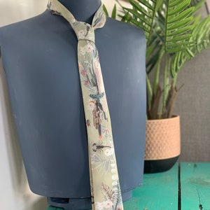 May include: A beige silk tie with a floral and insect print. The tie has a pattern of white flowers, green leaves, and brown insects. The tie is tied around a gray mannequin.