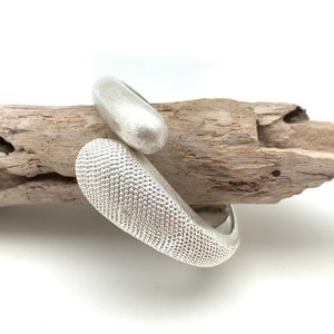 Puede incluir: Un brazalete de plata con un diseño ovalado texturizado. El brazalete está hecho de dos piezas que se unen.