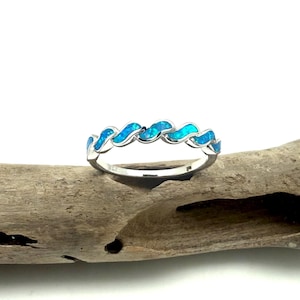 Puede incluir: Un anillo de plata con un diseño ondulado, con secciones alternas de piedras de ópalo azul brillante. El anillo se encuentra sobre un fondo de madera natural, creando un contraste de texturas y colores. El diseño del anillo es elegante y llamativo.