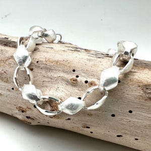 May include: A silver bracelet with a textured finish. The bracelet features a series of oval and diamond-shaped links. The jewelry is displayed on a piece of driftwood.