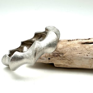 May include: A silver bracelet with a textured, organic design. The bracelet is resting on a piece of driftwood.