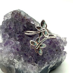 May include: A silver ring with a double dragonfly design, set against a purple amethyst crystal. The dragonfly wings have a blue-green tint. The ring's design is delicate and detailed, showcasing a nature-inspired theme.