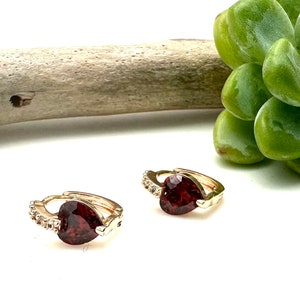 Pode incluir: Um par de brincos de argola de ouro com uma pedra vermelha em forma de coração e detalhes de cristal transparente.