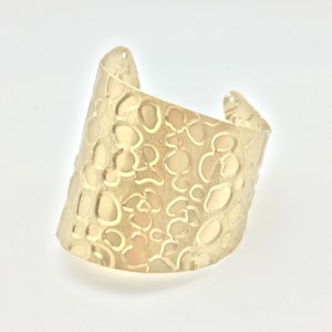 May include: A gold-toned cuff bracelet with a textured surface featuring a repeating pattern of small circles and ovals.
