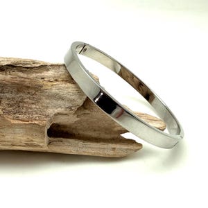 May include: A silver-colored bangle bracelet with a smooth, polished surface. The bracelet is round and sits on a piece of driftwood against a white background. The bangle is approximately 2.5 inches in diameter.
