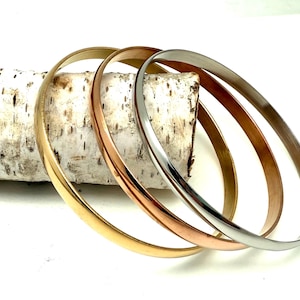 Può includere: Quattro braccialetti in metallo dorato, oro rosa e argento riposano su un pezzo di corteccia di betulla. I braccialetti sono lisci e lucenti, riflettendo la luce. Un set di gioielli semplice ed elegante.