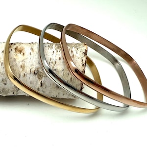 May include: Three square-shaped bangles in gold, silver, and rose gold rest on a piece of birch bark. The bangles have a smooth, polished finish and a minimalist design. The gold bangle is on the bottom, followed by silver and rose gold.