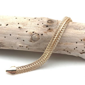 May include: A gold chain bracelet with a woven design. The bracelet is lying on a piece of driftwood.