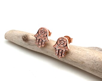 May include: A pair of rose gold earrings featuring a hamsa hand design. Each earring is set with clear cubic zirconia stones.