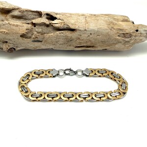 May include: A stainless steel and gold-tone bracelet with a chain link design. The bracelet is displayed on a white surface with a piece of driftwood in the background. The bracelet has a clasp closure.