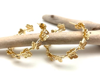 May include: Gold-colored hoop earrings with a butterfly design. The earrings are adorned with multiple butterfly shapes along the hoop's circumference. The earrings are displayed on a piece of driftwood against a white background.