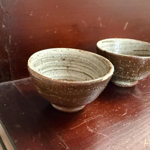 Puede incluir: Dos cuencos de cerámica hechos a mano con una estética rústica. Los cuencos tienen un exterior marrón y un interior color crema. Los cuencos están colocados sobre una superficie de madera oscura.