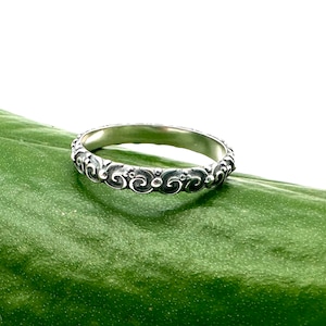 Peut inclure: Une bague en argent avec un motif délicat et complexe de volutes. La bague est posée sur une feuille verte.