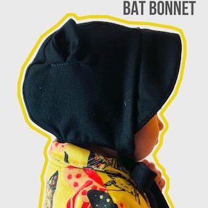 May include: A black 100% wool bat bonnet with ear flaps. The bonnet is worn by a child wearing a yellow and red patterned shirt.