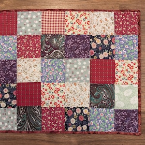May include: A patchwork quilt square with a variety of fabric patterns. The square is made up of smaller squares in shades of red, purple, green, and white. Patterns include polka dots, floral prints, and paisley designs. The quilt has a red border.