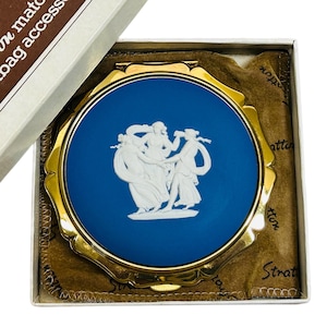 May include: A vintage Stratton compact mirror in its original box. The compact features a blue base with a white relief design of three figures. The gold-toned frame and the box are visible.