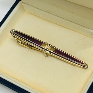 May include: A gold-toned tie clip with a burgundy enamel center and decorative emblem, presented in a blue velvet lined box. The clip is rectangular and has a clasp on the back.
