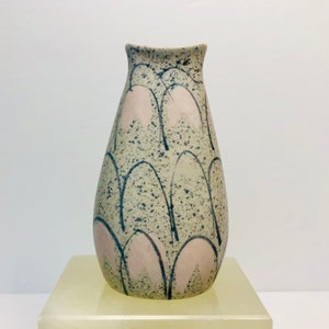 May include: A small, light grey ceramic vase with a speckled glaze. The vase has a rounded, teardrop shape and is decorated with a pattern of pink and black arches.