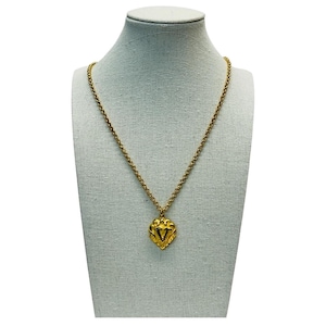 May include: A gold-toned necklace with a heart-shaped pendant. The pendant features a decorative design with a central V-shaped element. The necklace is displayed on a mannequin.