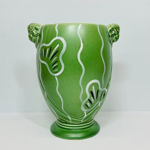 May include: A green ceramic vase with white and black stylized floral designs and ram's head handles.