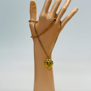 May include: A gold-colored necklace with a heart-shaped pendant. The pendant features a decorative design with a central V-shape. The necklace is displayed on a mannequin hand against a white background. The chain is approximately 18 inches long.