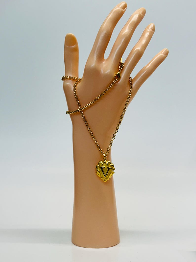 May include: A gold-colored necklace with a heart-shaped pendant. The pendant features a decorative design with a central V-shape. The necklace is displayed on a mannequin hand against a white background. The chain is approximately 18 inches long.