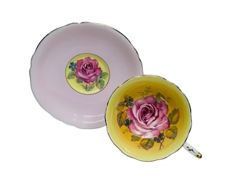 May include: A vintage teacup and saucer set. The saucer is light pink with a central floral design. The teacup interior is yellow with a large pink rose. Both pieces have gold trim.