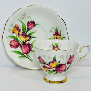 May include: A white teacup and saucer with a floral design featuring pink and yellow tulips and white flowers. The edges of the cup and saucer are trimmed with gold.