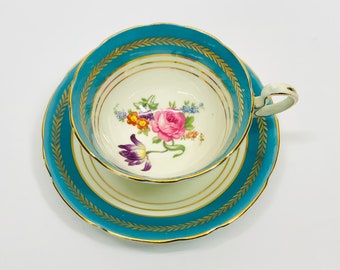May include: A vintage teacup and saucer set with a floral design. The cup and saucer are both a light blue color with a gold trim. The floral design features a pink rose, purple tulips, and other colorful flowers.