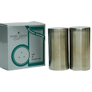 Puede incluir: Un juego de salero y pimentero de Georg Jensen, presentado en una caja. El juego incluye dos saleros y pimenteros cilíndricos de metal cepillado. La caja es blanca con un borde verde y el texto "Season's Greetings".