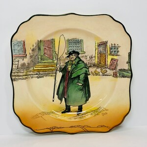May include: A vintage ceramic plate with a colorful illustration of a man in a green coat and hat holding a stick, walking down a street with buildings on either side. The plate has a scalloped edge and a brown rim.