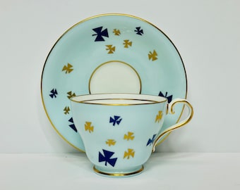 May include: A light blue teacup and saucer set with a gold rim. The cup and saucer are decorated with a pattern of blue and gold shamrocks.