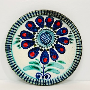 May include: A white ceramic plate with a blue and red floral design. The flower has a blue center with a white grid pattern and red dots around the edge. The flower is surrounded by green leaves.