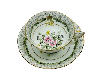 Könnte beinhalten: Eine weiße Teetasse und Untertasse mit floralem Design und goldener Verzierung. Die Tasse zeigt eine Krone und die Buchstaben "ER" in Gold.
