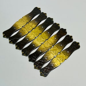 Op de afbeelding: Acht antieke metalen ladegrepen met een zwarte en gouden afwerking. De grepen hebben een decoratief, sierlijk ontwerp.