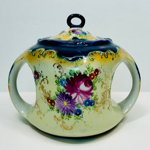 May include: A white porcelain teapot with a blue and gold floral design. The teapot has two handles and a lid with a knob on top.