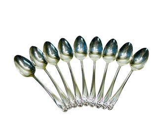 May include: A collection of nine silver spoons arranged in a fan shape. Each spoon has a polished, oval bowl and a slender handle with a decorative floral design near the base. The spoons are set against a white background.
