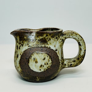 May include: A small, brown and white ceramic creamer with a speckled glaze and a rounded handle.
