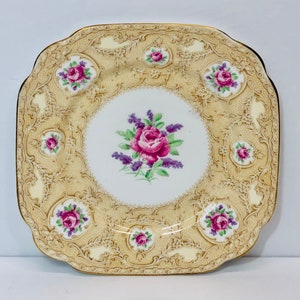 Op de afbeelding: Een vierkant, crèmekleurig keramisch bord met een goudkleurig reliëfdesign. Het midden van het bord heeft een roze en paarse roos met groene bladeren en paarse bloemen.