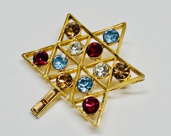May include: A gold-toned star of David brooch with a geometric design and colorful rhinestones. The rhinestones are in shades of red, blue, and brown.