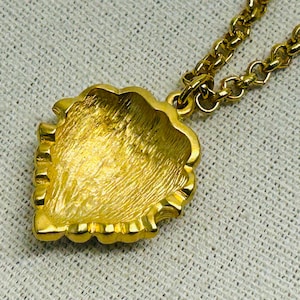 May include: A gold-toned necklace featuring a pendant with a textured, leaf-like design. The pendant is intricately detailed and suspended from a matching gold chain. The necklace is displayed on a textured, off-white surface, highlighting the jewelry's shine.