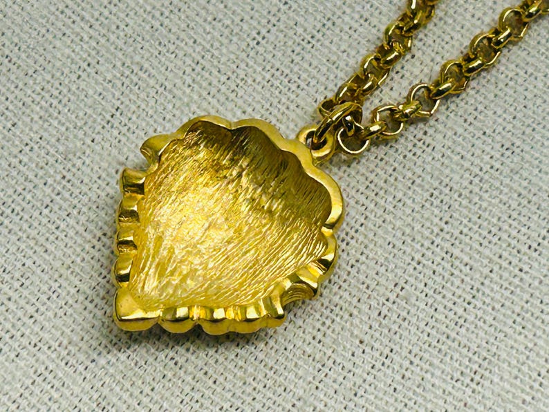 May include: A gold-toned necklace featuring a pendant with a textured, leaf-like design. The pendant is intricately detailed and suspended from a matching gold chain. The necklace is displayed on a textured, off-white surface, highlighting the jewelry's shine.