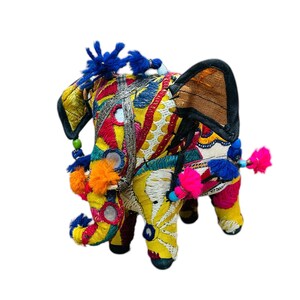 May include: A colorful, embroidered elephant figurine. The elephant is adorned with vibrant yellow, red, and blue fabrics, accented with mirrors and tassels. The ears are a tan color with black trim. The trunk and tail feature pink and blue tassels.