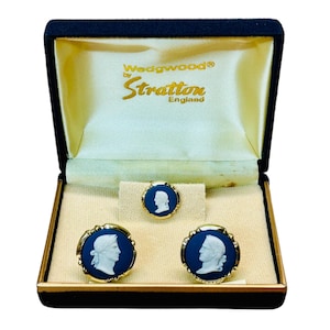 Op de afbeelding: Een vintage sieradenset van Wedgwood by Stratton England, gepresenteerd in een zwarte doos met gouden randen. De set bevat manchetknopen en een stropdaspen, elk met een blauw camee-ontwerp en een wit profiel. De binnenkant van de doos is gevoerd met crèmekleurig satijn.