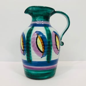 May include: A green ceramic pitcher with a white background and colorful abstract designs. The pitcher has a wide mouth and a handle.